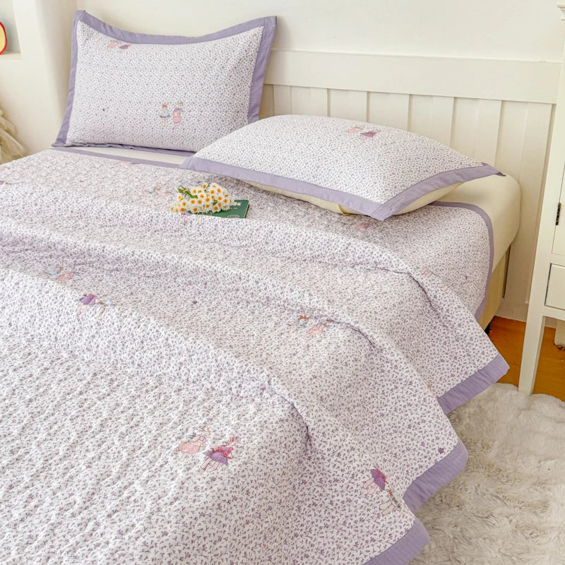 A light purple floral quilted bedspread with pink and white patterns is displayed on a bed.
