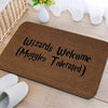 A brown entrance mat with black text reading Wizards Muggles Welcome Tolerated placed on a wooden surface.