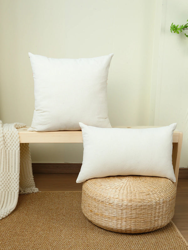 A white cushion cover with elegant floral embroidery resting on a wooden bench next to a woven stool.
