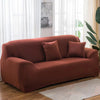 A modern, rust-colored stretch sofa cover with two throw pillows placed in a bright living room.