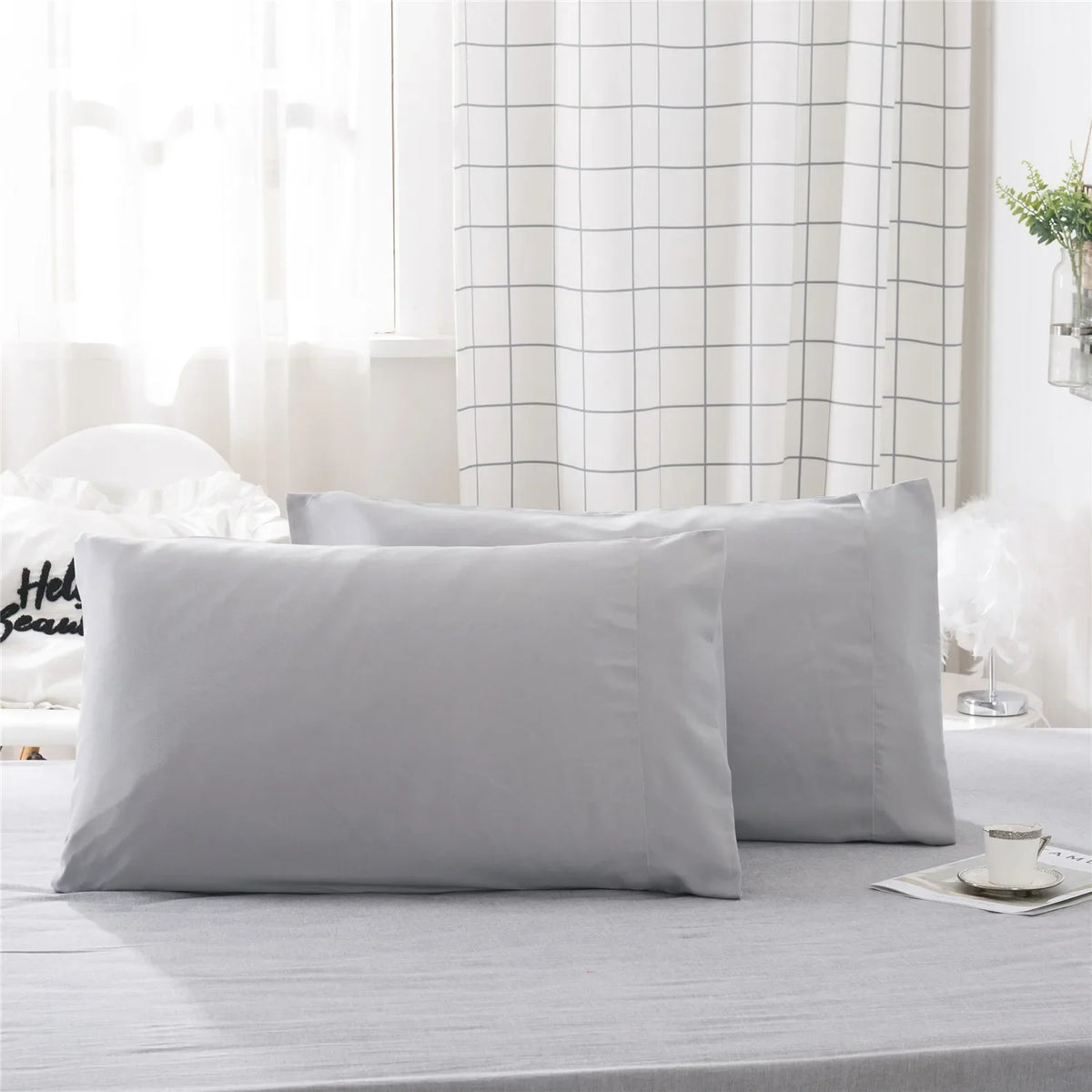 A set of two light gray soft polyester pillow covers displayed on a bed with a white checkered curtain backdrop.