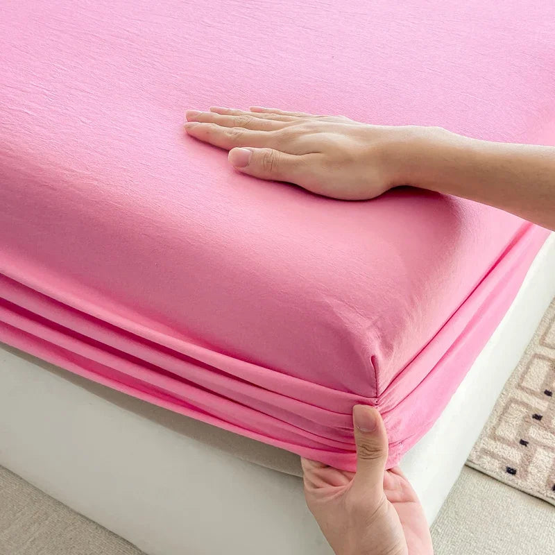 A hand is folding a bright pink fitted bed sheet made of premium 100% cotton.