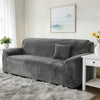 A charcoal gray stretch velvet sofa slipcover with a throw pillow on a light rug in a bright room.