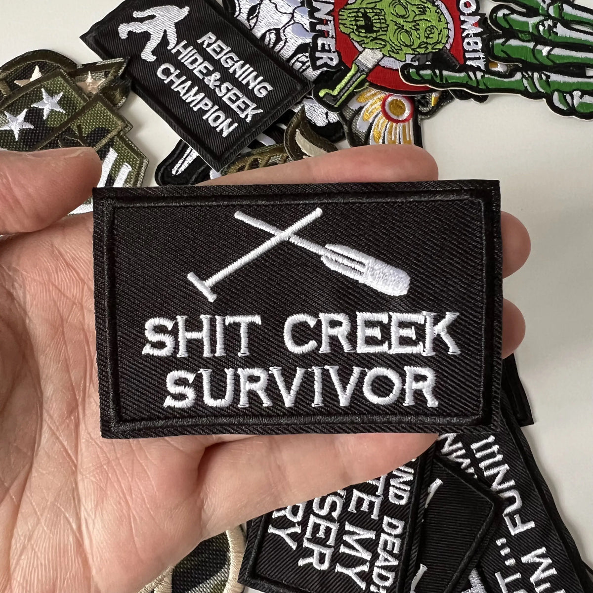A black iron-on patch with white text reading SHIT CREEK SURVIVOR is held in a hand among other patches.