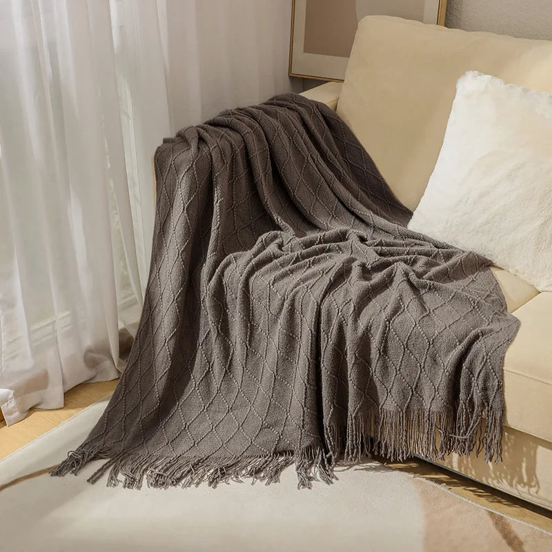 A dark gray oversized knitted tassel throw blanket draped over a light-colored sofa.