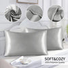 A silver satin pillowcase is displayed next to a white wooden headboard and a cup, highlighting its soft and cozy texture.