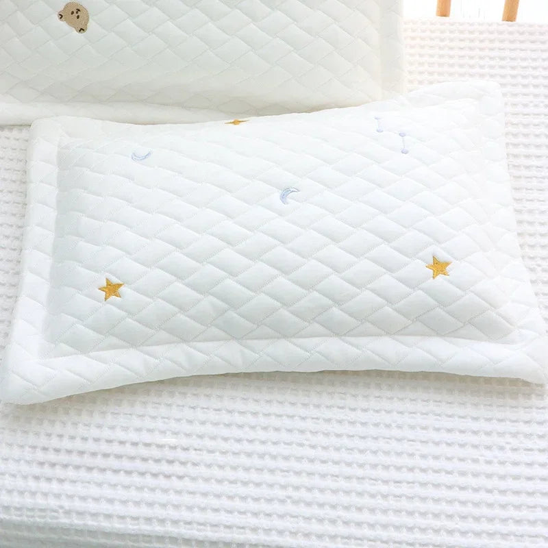 A white quilted pillow with gold stars and blue crescent moons resting on a white textured surface.