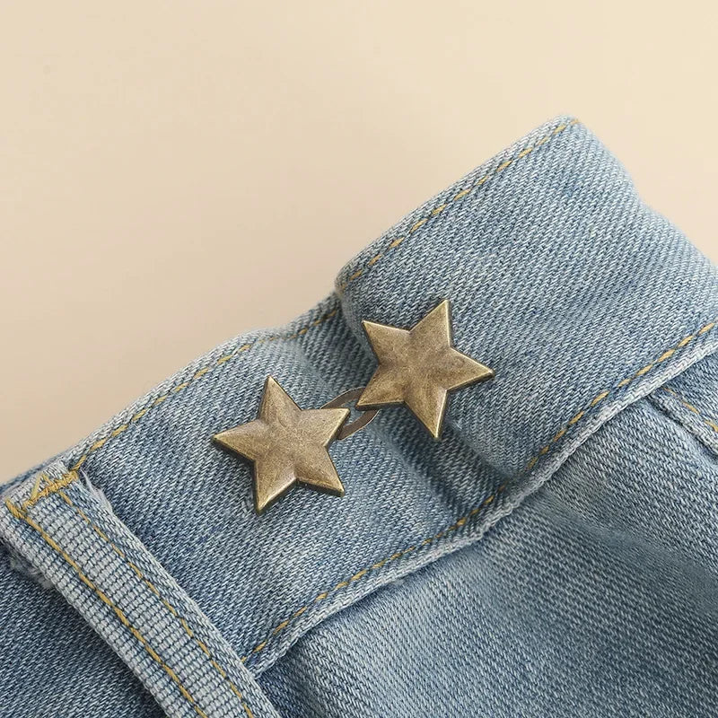 A close-up of blue denim fabric with two gold star-shaped button clips used for waist tightening.
