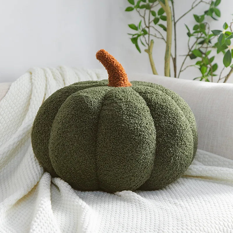 A round, moss-green plush pumpkin decoration with an orange stem resting on a white knitted fabric.