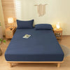 A dark blue fitted sheet set with matching pillowcases is displayed on a wooden bed frame in a bright bedroom.