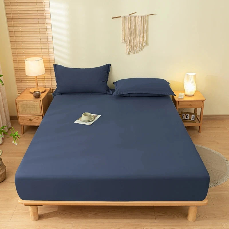 A dark blue fitted sheet set with matching pillowcases is displayed on a wooden bed frame in a bright bedroom.