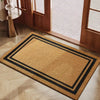 A rectangular natural coconut fiber doormat with black geometric borders placed on a light wooden floor.