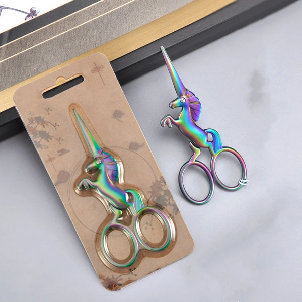 A pair of iridescent, three-color stainless steel unicorn sewing scissors resting on a kraft paper tag.