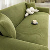 A moss green stretch jacquard sofa slipcover with a cream knitted throw pillow and a cup on the seat.