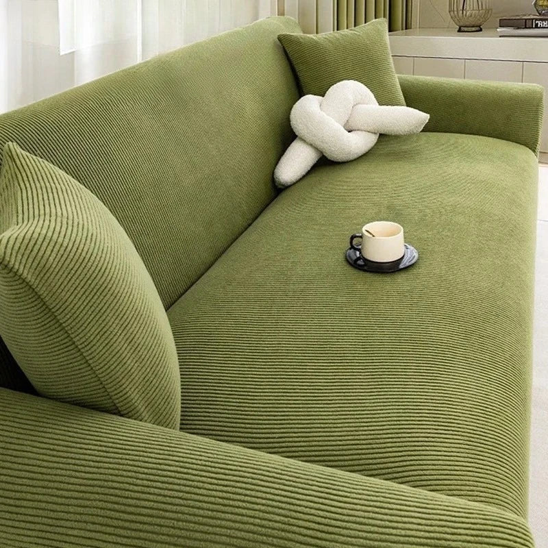 A moss green stretch jacquard sofa slipcover with a cream knitted throw pillow and a cup on the seat.