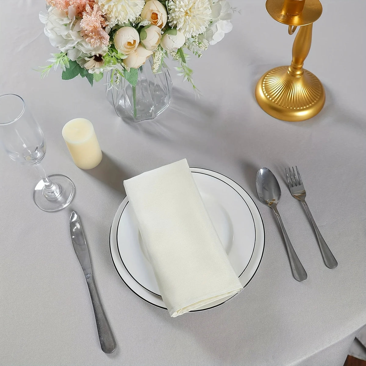A set of silky satin dinner napkins is displayed on a table with a gold candlestick and floral arrangement.
