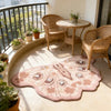 A round, irregular shaped plush floor rug in pink and beige tones sits on a light-colored floor near wicker chairs and a balc