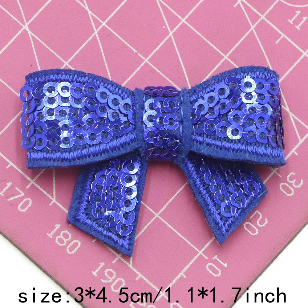A royal blue sequin bow patch for DIY decoration shown on a pink grid background.
