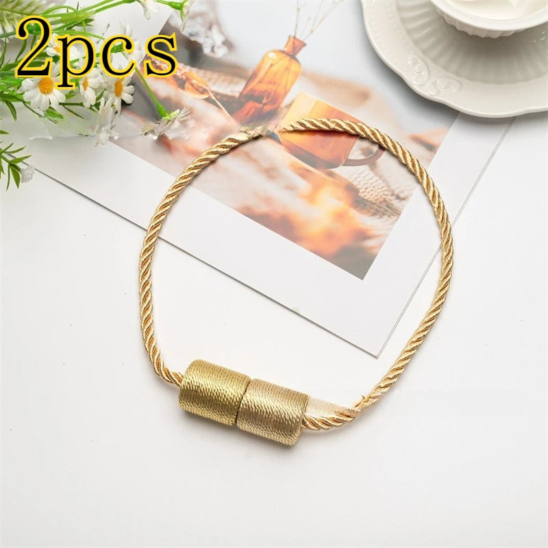 A set of two gold-colored magnetic curtain tiebacks with braided cord attached, displayed on a white surface.