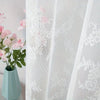 White lace curtain panel with floral embroidery and a pink flower arrangement in a light blue vase