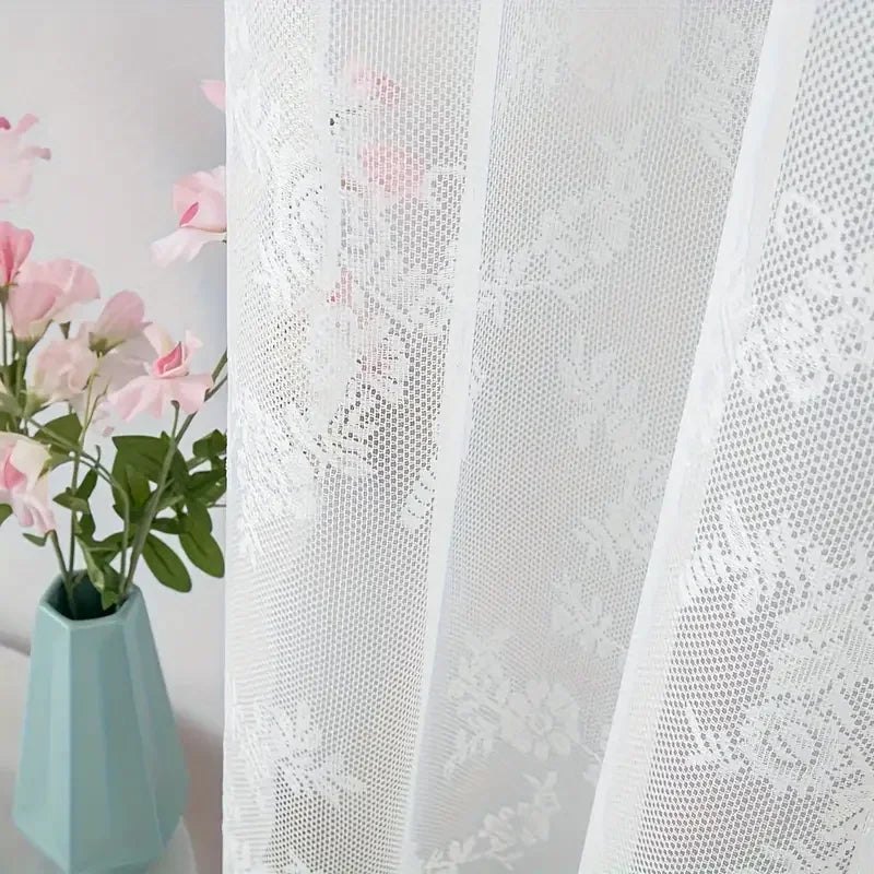 White lace curtain panel with floral embroidery and a pink flower arrangement in a light blue vase