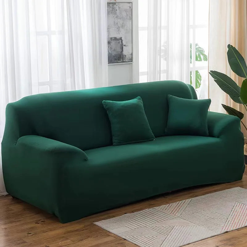 A deep emerald green stretch sofa cover with two accent pillows placed on a wooden floor near a window.
