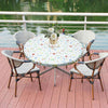 A round elastic-edged vinyl tablecloth set for outdoor dining with matching woven chairs by a body of water.