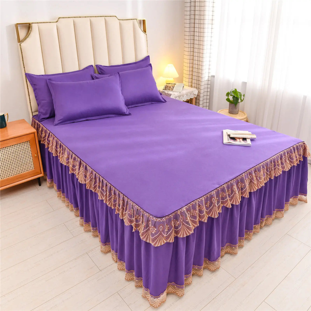 Purple luxury floral bed skirt protecting a mattress with a padded headboard and white curtains.