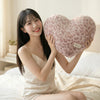 A young woman holds a pink leopard print plush heart-shaped pillow in a bright bedroom setting.