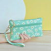 A hand holds a mint green PU leather coin purse with a floral pattern and a gold wrist strap against a yellow background.