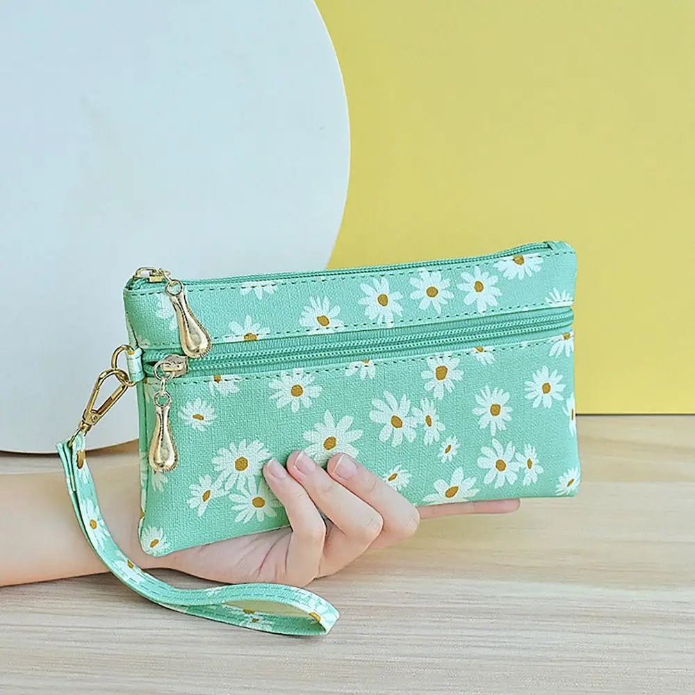 A hand holds a mint green PU leather coin purse with a floral pattern and a gold wrist strap against a yellow background.