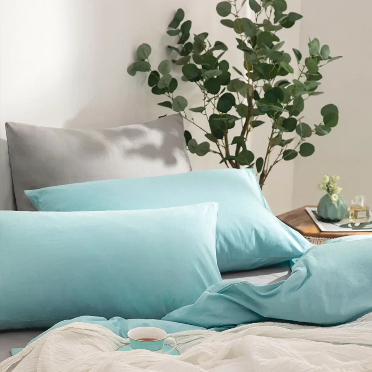 A set of four light blue microfiber pillow covers displayed on a bed with green foliage in the background.