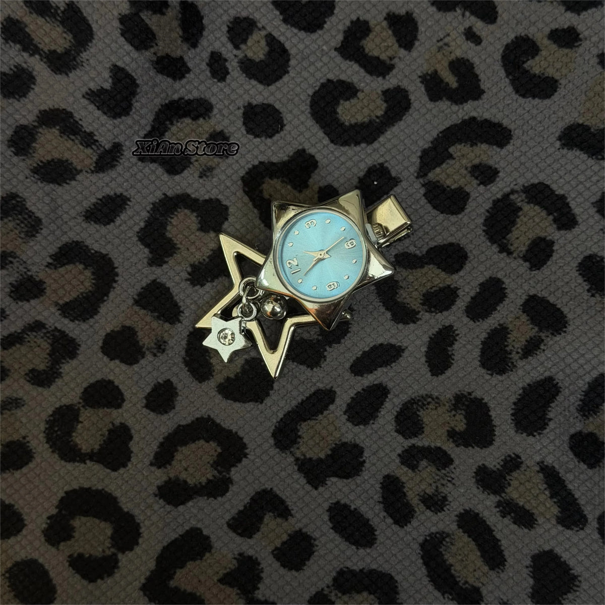 A silver star-shaped pocket watch keychain with a light blue face and hands is displayed on a leopard print background.