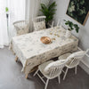 A dining table set with a botanical print linen tablecloth, white wooden chairs, and greenery in a bright room.