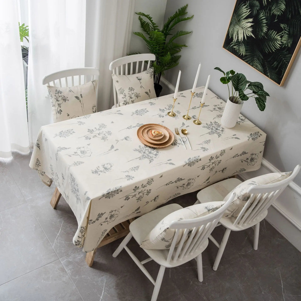 A dining table set with a botanical print linen tablecloth, white wooden chairs, and greenery in a bright room.