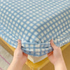 Hands are folding a light blue and white checkered flannel fitted sheet with deep pockets.