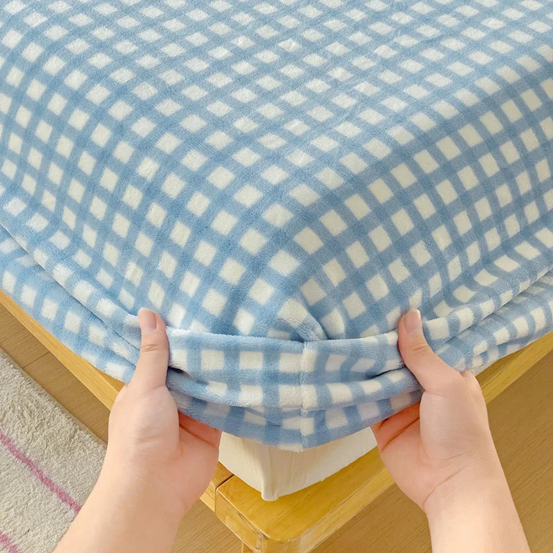 Hands are folding a light blue and white checkered flannel fitted sheet with deep pockets.