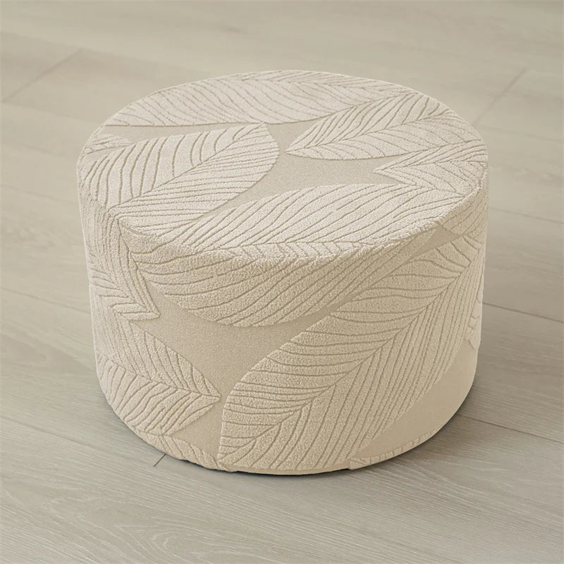 A round ottoman footrest slipcover in beige fabric featuring a woven leaf pattern on a light wood floor.