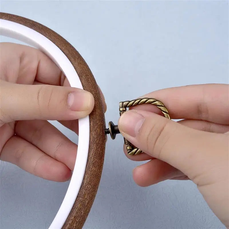 Hands are holding a round wooden embroidery hoop with a white cord and a gold-colored metal tool.
