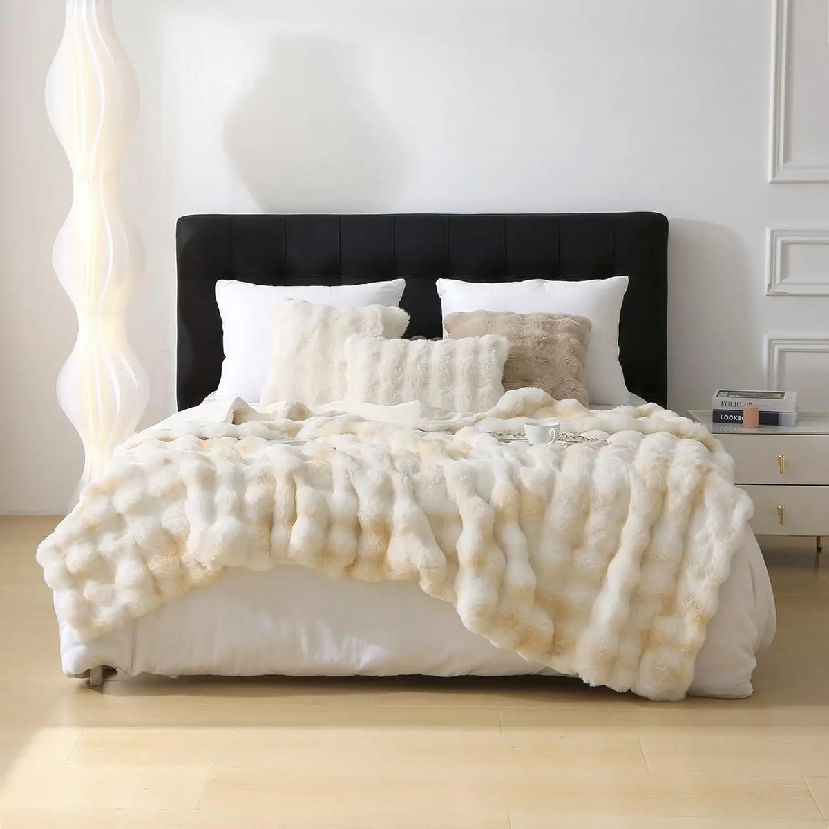 A luxury faux rabbit fur throw blanket draped over a white bed with black headboard and white pillows.
