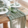 A rustic wooden table set with a sage green sheer gauze table runner, white plates, and silverware.