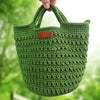 A hand-knitted green cotton yarn bag with a woven texture is being held against a blurred green outdoor background.
