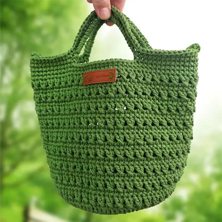 A hand-knitted green cotton yarn bag with a woven texture is being held against a blurred green outdoor background.