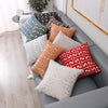 A collection of geometric patterned lumbar pillows in shades of orange, gray, and white on a gray sofa.