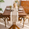 A rustic wooden table set with a beige macrame tassel table runner, ceramic vessels, and natural decor.