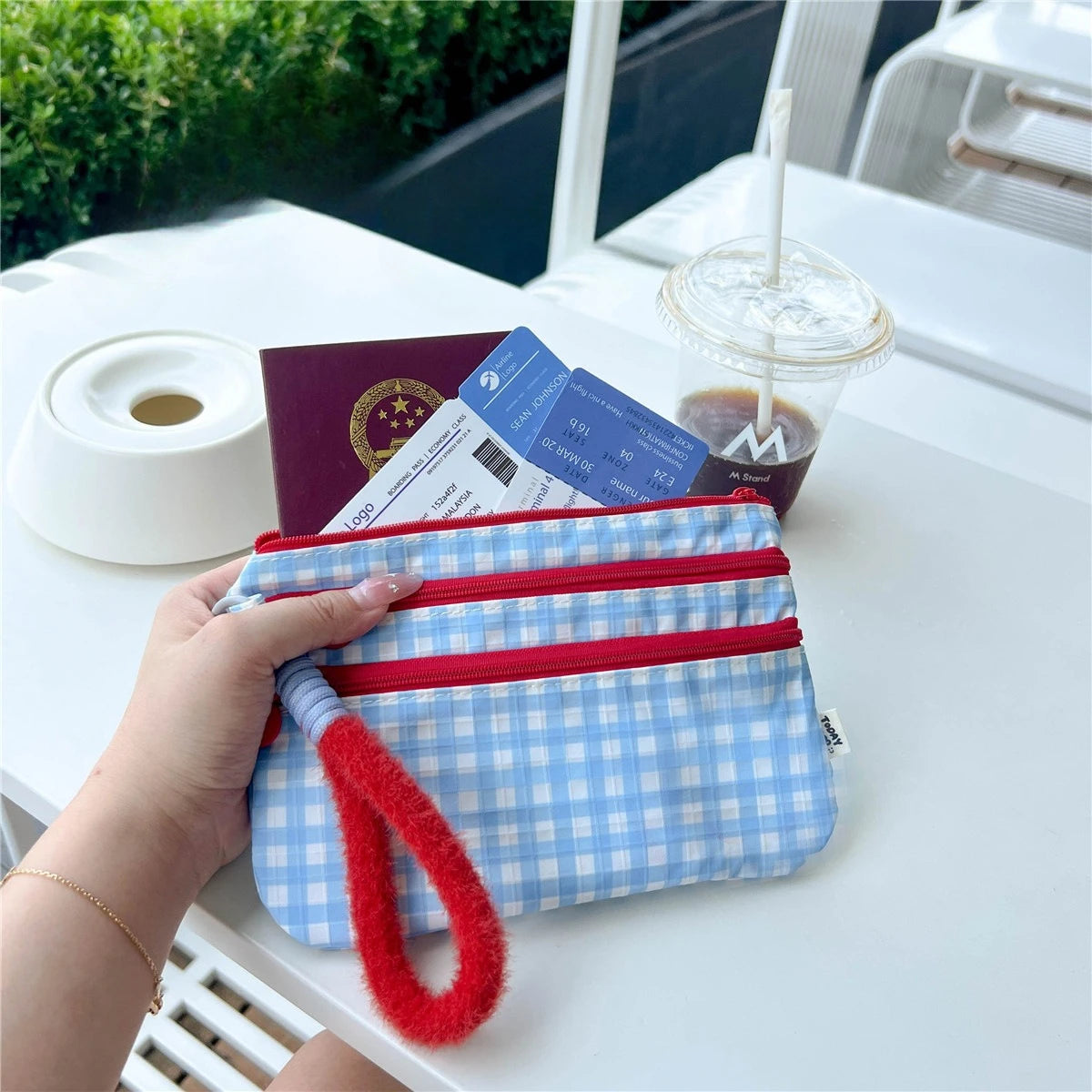 A hand holds a blue and white plaid waterproof passport holder next to a coffee cup and ID cards on a white table.