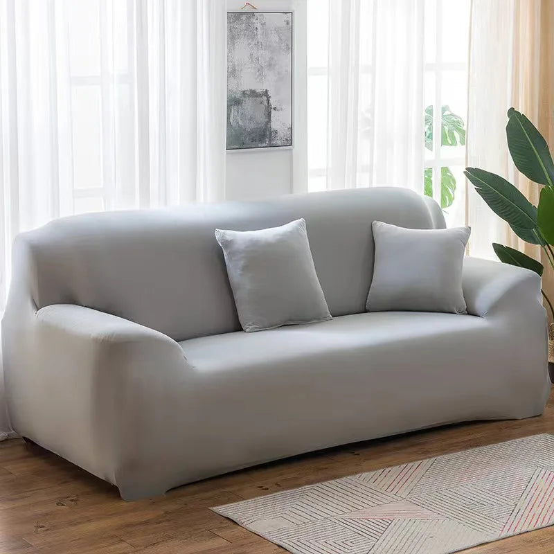 A light gray stretch sofa cover placed on a wooden floor near a window.