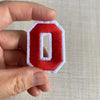 A hand holds a red felt number patch shaped like the letter O with white stitching.
