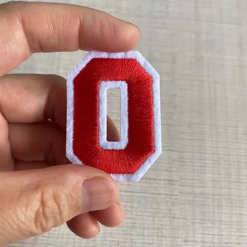 A hand holds a red felt number patch shaped like the letter O with white stitching.