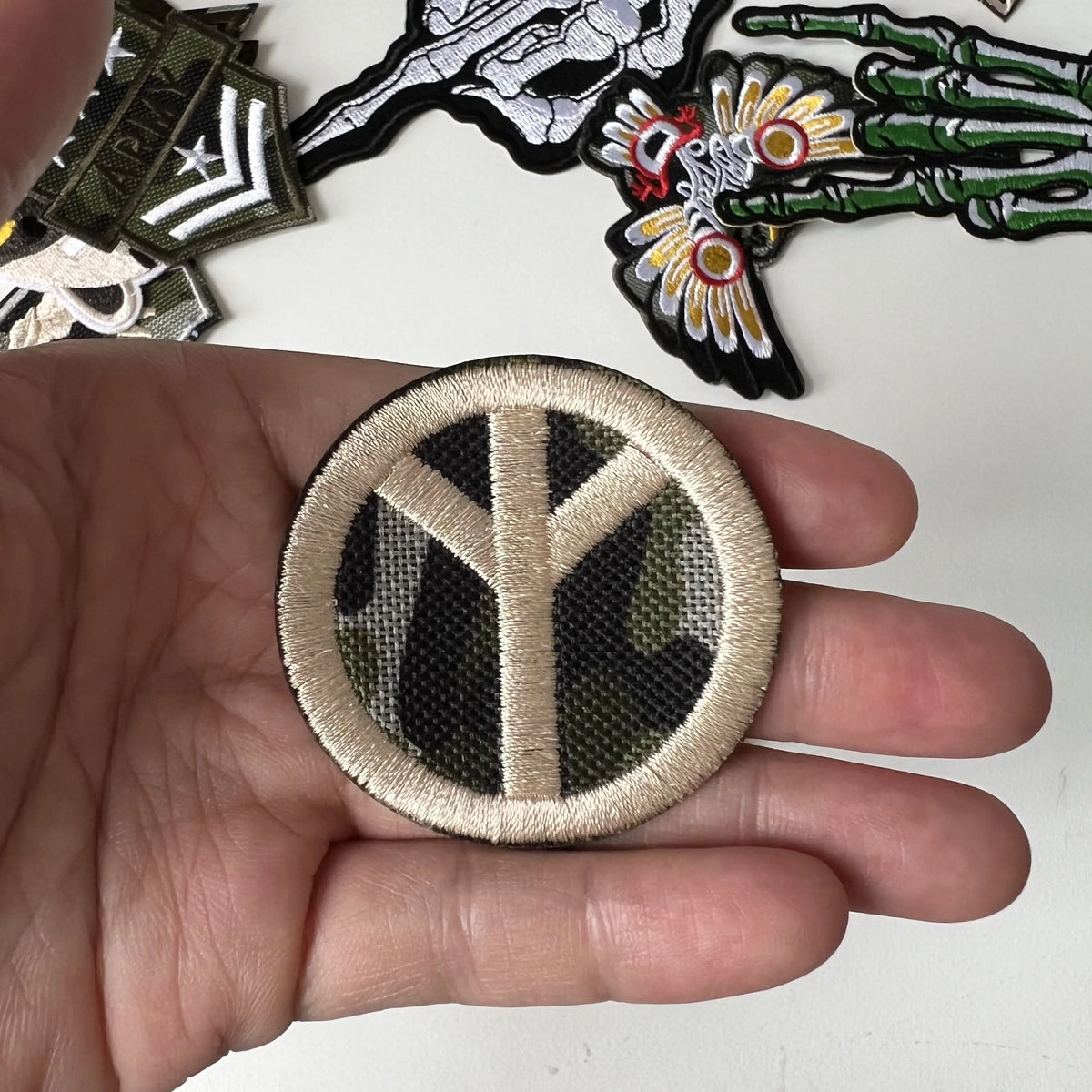 A hand holds a round embroidered patch featuring a stylized tree design in olive green and beige tones.